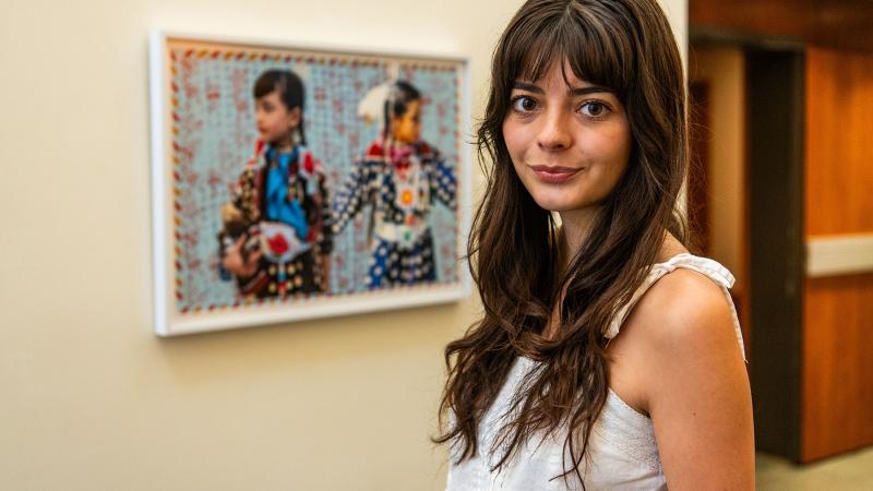 Julia Pippenger with long dark hair and wearing a white strappy shirt, standing next to Indigenous artwork of two young people. 