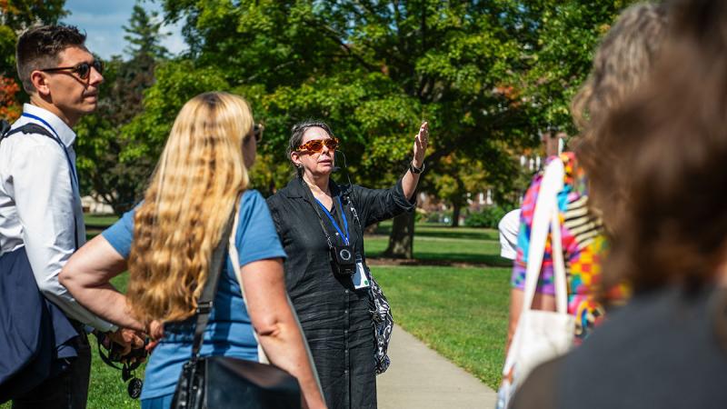 Person with rose colored glasses standing outside in the Vassar Quad speaking with their hand raise while giving a tour with others looking on.