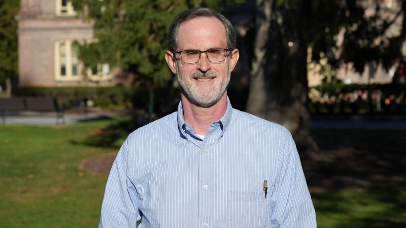 Ken Foster wearing a light blue with white striped collared shirt, eyeglasses, and short hair with beard and mustache with a brick building and large tree in the background.