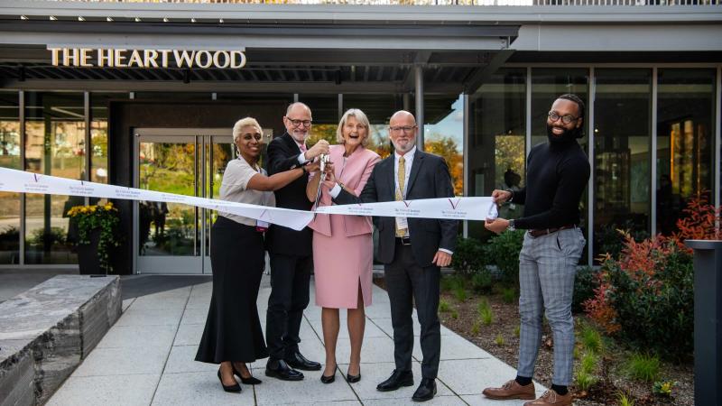 Five smiling people standing in front of a building all participation in cutting a ceremonial ribbon.