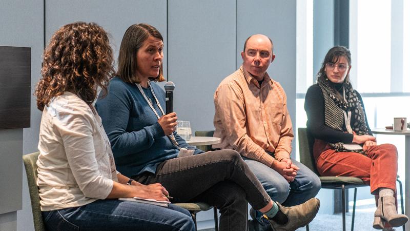 Panel of three people sitting on a stage with one person speaking into a microphone while the others look on.