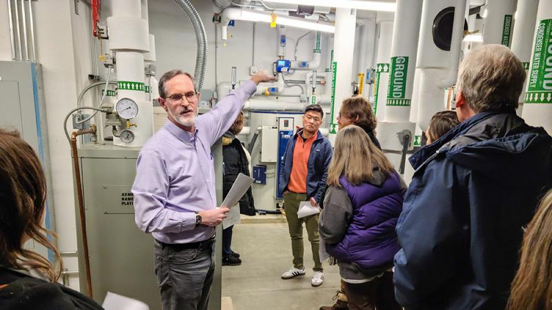 A person in a light blue button-up collared shirt lecturing to an audience while standing in a well lit mechanical room with pipes and systems in the background.