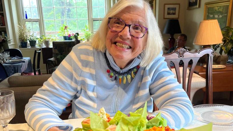 A photo of Marian Heath Mundy, a person with long gray hair, glasses, and a blue shirt. Mundy is sitting at a table with flowers on it.