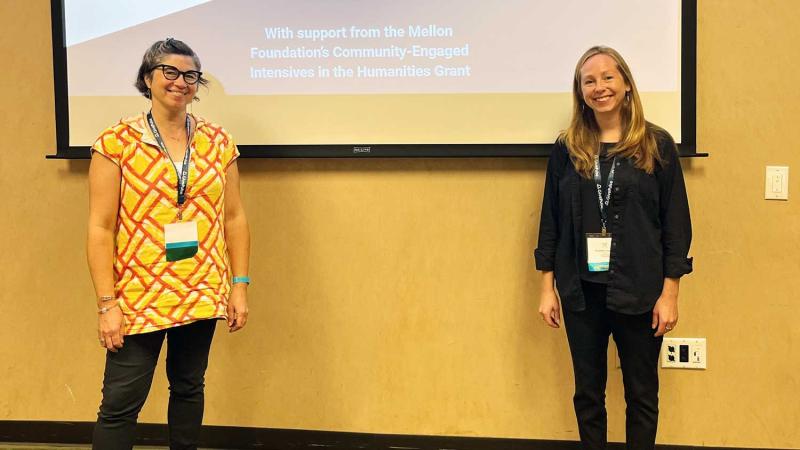 Two smiling people standing in front of a projected slide presentation on a wall.