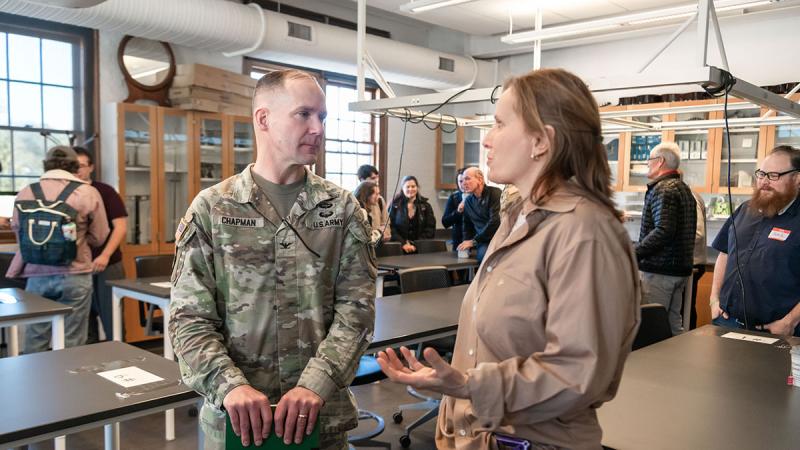 Two people standing in a classroom speaking to each other with other people and classroom tables in the background. One of the people speaking is wearing a camouflage, military uniform. 