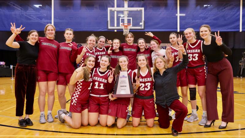 Basketball team and coaches making victory signs with their hand and posing on the court with a trophy.