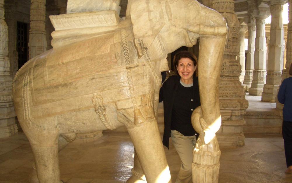 A person with short dark hair, a dark sweater, and beige pants is standing beneath the trunk of a stone-carved elephant smiling at the viewer.