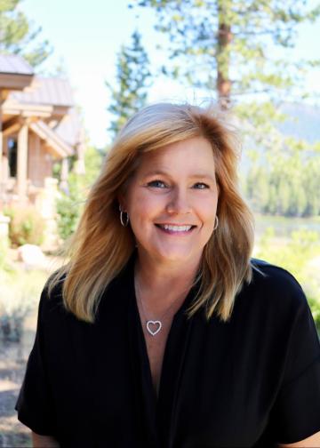 A person with long blond hair and a dark shirt smiles at the viewer. The person is outside on a sunny day, standing in front of trees and a house.