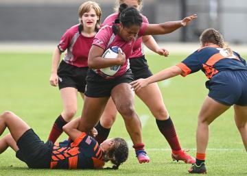 Person holding a rugby ball while others fight and struggle to stop, grab, hold her while playing rugby on a field.