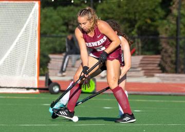 Person in a Vassar maroon uniform holding a field-hockey stick preparing to hit the ball on a green field with a net in the background.