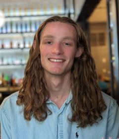 Person with long brown hair and a blue button shirt sitting in a lounge with bottles on the wall in the background.