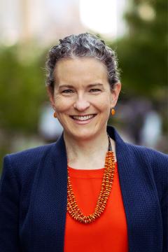 Person wearing a orange shirt, blue jacket, orange beaded necklace, round orange earrings, and has short hair. 