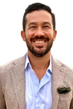 Person wearing a light blue button down collared shirt, light tan jacket, with short dark hair, beard and mustache. 