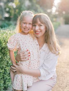 Amanda Zeligs Hand wearing white pants and shirt and has long light hair squatting and holding a young girl who is wearing a white dress with red and pink hearts. 