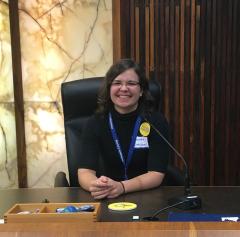 Chelsea Sheldon wearing a black shirt with name sticker, sitting in a black chair at a desk with hands clasped on it. 