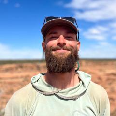 Ethan Pierce wearing a light blue shirt, cap with sunglasses on top and a brown beard. 