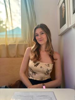 Olivia Lerman wearing black pants and beige and black top sitting in a chair in front of a table.