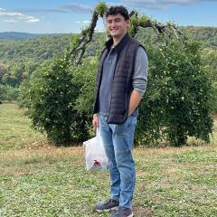 Ryan Mazurkiewicz wearing a blue shirt, blue jacket vest, jeans while standing in front of a tree. 