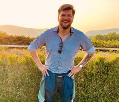 Sam Rebelein wearing a blue and white checkered shirt, jeans, and a jacket tied around his waist and has short light hair and beard with hands on hips and green foliage in the background.