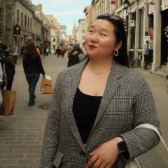 Sophia Wood wearing a black shirt, striped patterned jacket, looking to the side while in a street with people and shops.