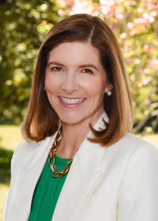 A person with long brown hair and a white format jacket smiles at the viewer while standing in front of foliage on a sunny day.