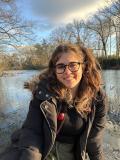 Person sitting outside with a lake in the background wearing a winter jacket. The person has long brown hair and glasses.