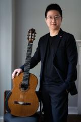 Person with a dark suit standing next to a classical guitar in a portrait.