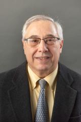 Portrait of Anthony Scaduto. Person with grey hair and glasses wearing a black suit and tie with a yellow shirt.