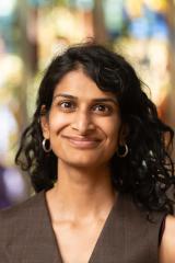 Portrait of a person with long black hair and olive brown blouse smiling with a blurry stained glass window in the background.