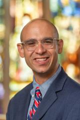 Portrait of a person in a blue suit and glasses in front of a stained glass window.