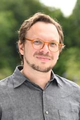 Portrait of a person outside with trees in the background wearing a grey, button-down, collared shirt with glasses and a thin mustache and beard.