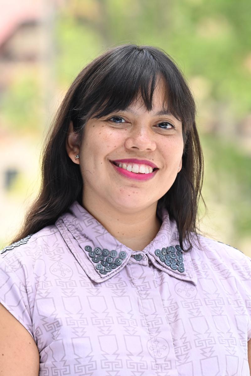 Portrait of a person in a lavender collared shirt with long dark hair standing outside with a vague, blurry building and trees in the background.