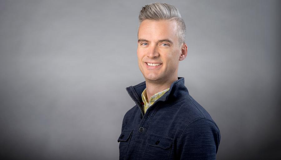 Colin Echeverría Aitken wearing navy shirt with high collar against a gray background.