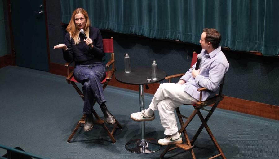 Two people seated in directors chairs on a stage holding microphones having a conversation.