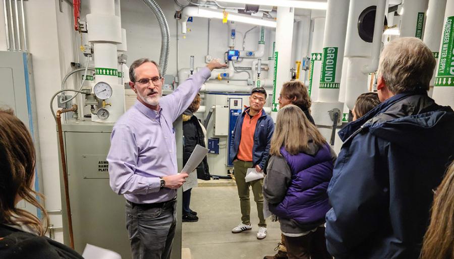 A person in a light blue button-up collared shirt lecturing to an audience while standing in a well lit mechanical room with pipes and systems in the background.