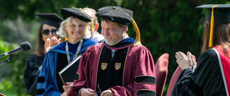 Smiling person in graduation ceremony attire receiving applause from others on stage.