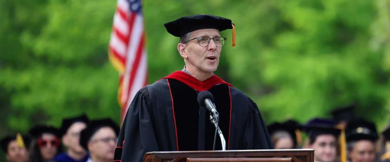 Person in graduation ceremony attire speaking on stage behind a microphone and a lectern.