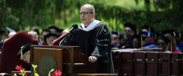 Person in graduation ceremony attire speaking on stage behind a microphone and a lectern.