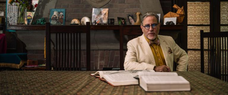 Person in a white suit seated behind a table behind two open books.