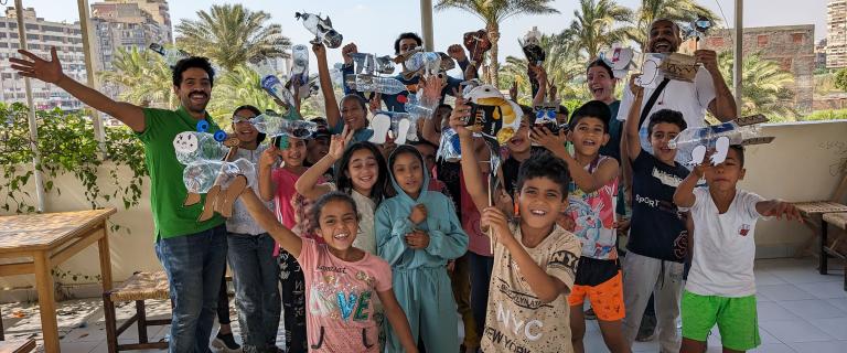 Many children standing in a group for a photo waving while standing outside under a tarp in Cairo, Egypt.