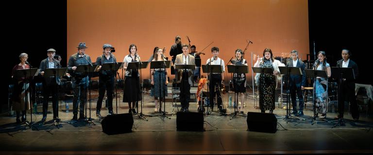 A large group of people sanding in a line on a dark stage in front of music stands singing.