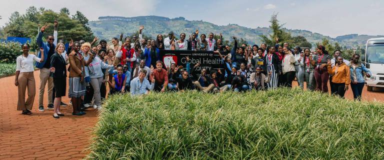 Large group of smiling waving people standing in front of a mountain scape and behind a sign that reads, University of Global Health.