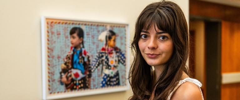 Julia Pippenger with long dark hair and wearing a white strappy shirt, standing next to Indigenous artwork of two young people. 