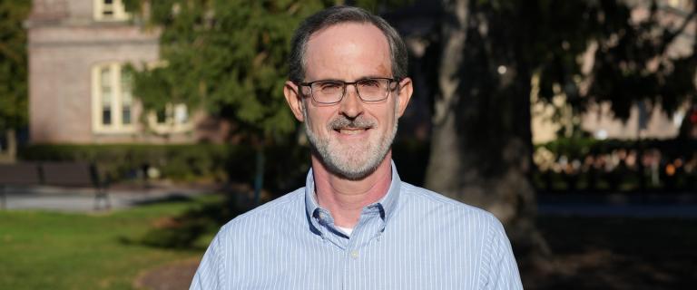 Ken Foster wearing a light blue with white striped collared shirt, eyeglasses, and short hair with beard and mustache with a brick building and large tree in the background.