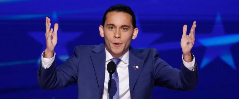 Person standing behind and lectern and microphone with both arms in the air speaking emphatically.