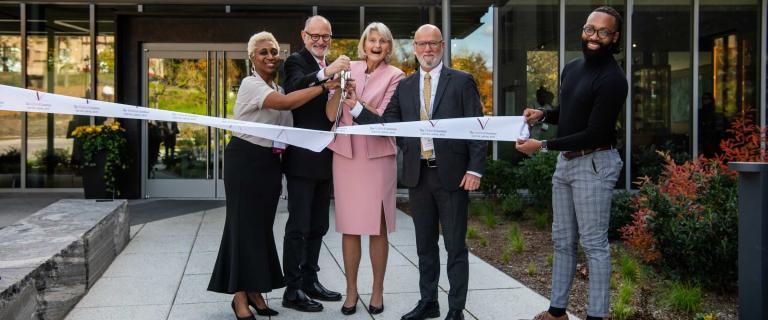 Five smiling people standing in front of a building all participation in cutting a ceremonial ribbon.