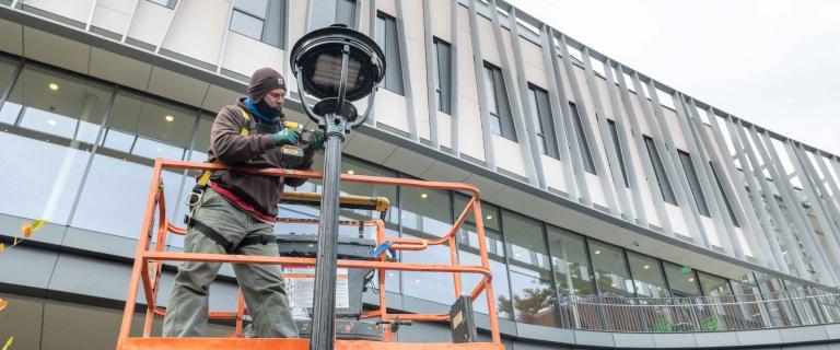 Person in a lift repairing a light with a glass and metal building in the background.