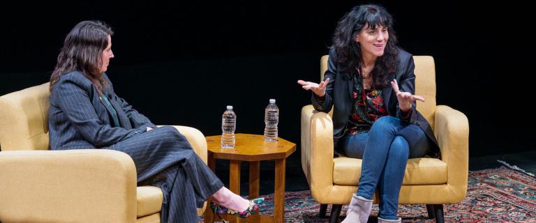 Two people sitting on a dark stage in large comfortable camel-tan chair speaking to each other and the audience.