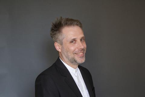 Person standing for a portrait with a black suit jacket and white collared shirt buttoned to the top.