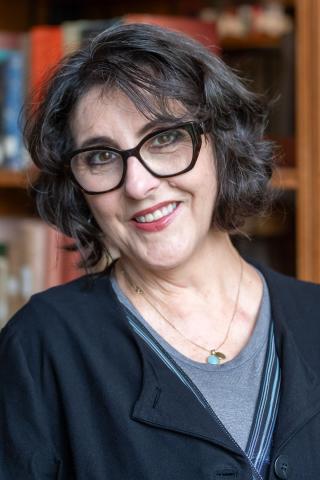 Kristin Sánchez Carter wearing a black jacket over a gray shirt with large bookcases in the background.